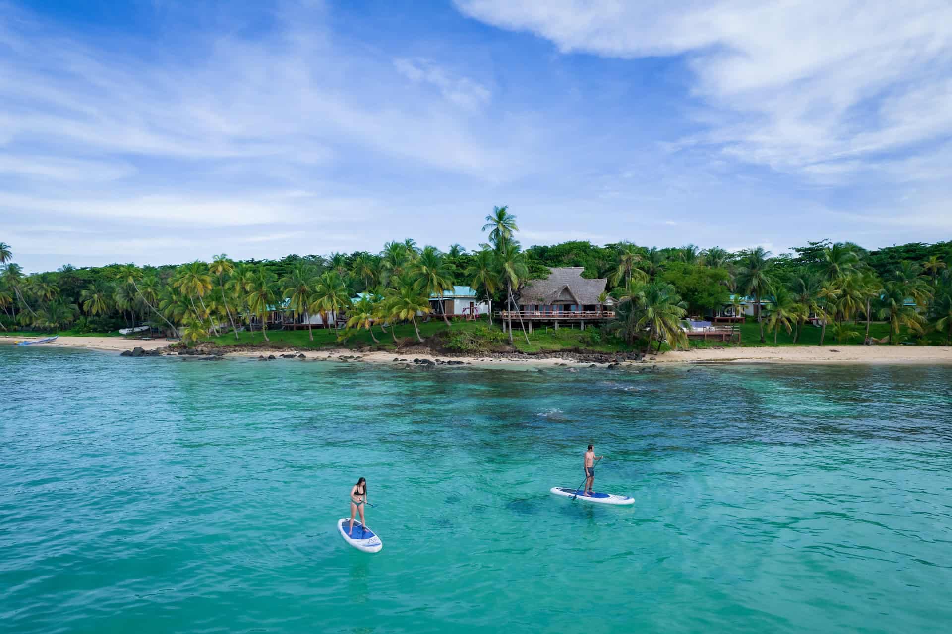 A woman and man paddle boarding over clear turqoise water in front of Yeyama Reef Resort on Little Corn Island, Nicaragua